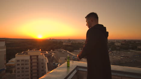 Close-the-lid-of-the-laptop-and-drink-beer-standing-on-the-roof.-A-man-in-a-hoodie-with-a-laptop-in-his-hands-at-sunset.